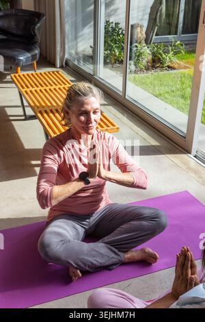 Senior friends practicing yoga on purple yoga mat in home living area with smartwatch on wrist. Wellness, mindfulness, relaxatio Stock Photo