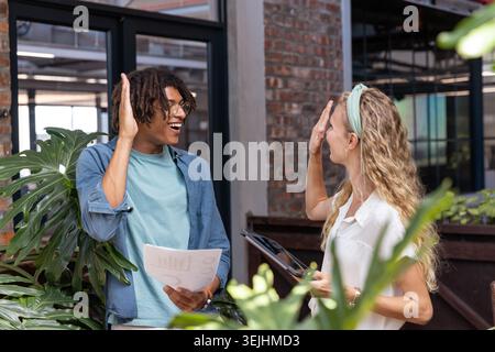 Diverse coworkers high-fiving while exchanging chart sheets and tablet in modern office with plants. Collaboration, teamwork, pr Stock Photo