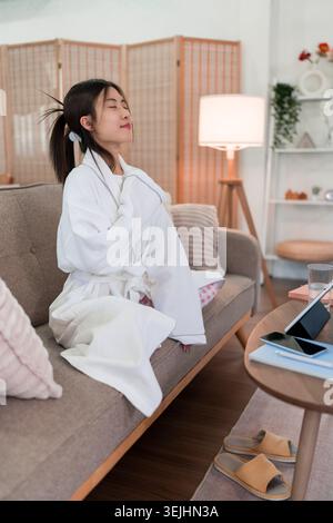 young Asian woman relaxing in white bathrobe combing hair after shower at modern home living room beauty wellness Stock Photo