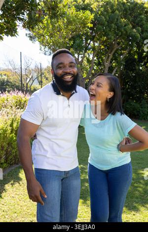 Diverse couple standing in sunlit garden on grassy lawn leaning toward each other laughing. Romantic, outdoor, leisure, natural, Stock Photo