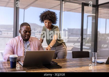 African American coworkers collaborating over laptop with coffee cup in glass-walled office. Collaboration, teamwork, modernity, Stock Photo