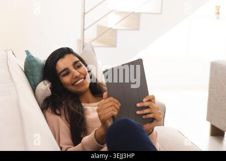 Asian woman smiling and holding tablet while reclining on sofa with teal cushion in living room. Modern, leisure, relaxation, te Stock Photo