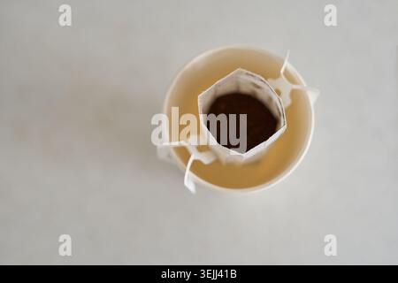 Overhead View Of Paper Cup With Drip Coffee Filter Filled With Grounds Inside Rim On Light Countertop Showing Brewing Setup And Clipped Paper Handles. Stock Photo