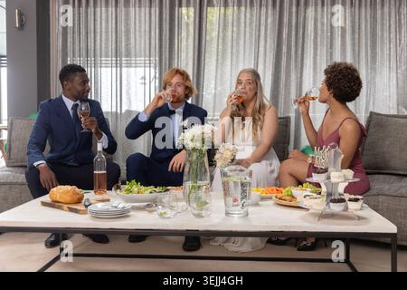 Diverse wedding party sitting on grey couch in lounge sipping rosé beside dessert stand. Elegance, celebration, camaraderie, so Stock Photo
