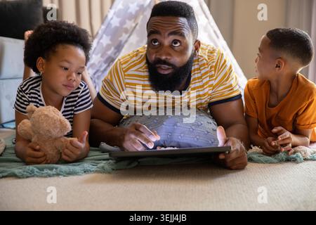 Diverse family lying on gray floor cushion at home using tablet with teddy bear near teepee. Family, parenting, relaxation, recr Stock Photo