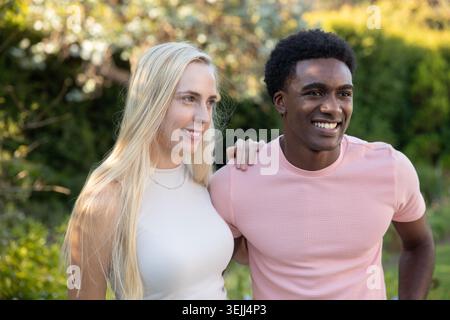 Diverse couple wearing sleeveless top and t-shirt standing in lush garden, smiling and gazing. Romance, harmony, serenity, outdo Stock Photo