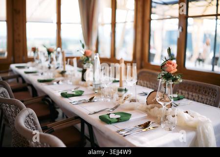 Elegant wedding reception table setting with white tablecloth, green napkins, candles, and fresh peach roses in a rustic wooden interior. Stock Photo