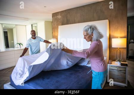 Couple making bed under white headboard in modern bedroom, spreading white bedsheet over mattress. Romantic, cozy, minimalist, i Stock Photo