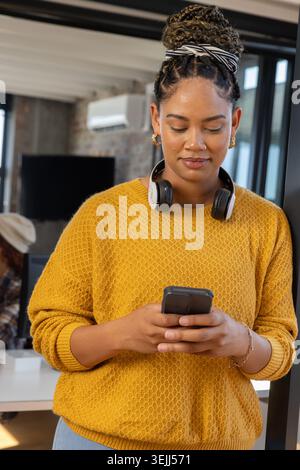 African American woman standing in modern office, checking smartphone with headphones and headband. Professional, workspace, con Stock Photo