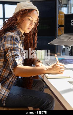 African American woman software developer sitting at office desk taking notes beside dual monitors. Technology, innovation, conc Stock Photo