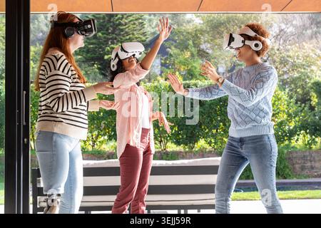 Diverse female friends interacting with virtual objects near bench on patio using VR headsets. Technology, innovation, collabora Stock Photo