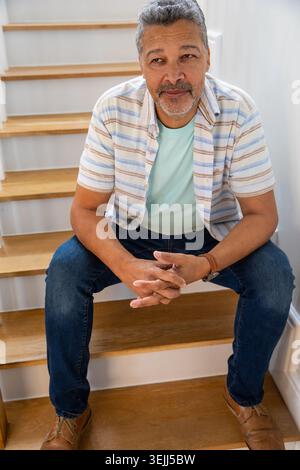 Senior Hispanic man leaning forward with clasped hands on wooden staircase at home. Elegance, contemplation, cozy, rustic, mature, lifestyle, relaxati Stock Photo