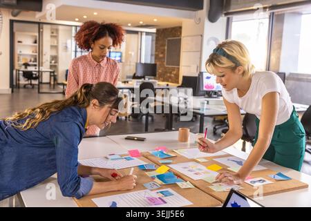 Diverse coworkers leaning over table in modern office arranging printed diagrams and sticky notes. Collaboration, teamwork, brainstorming, innovation, Stock Photo