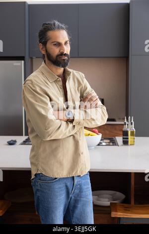 Mid adult Asian Indian man crossing arms in kitchen next to fruit bowl and oil bottles. Contemporary, lifestyle, minimalist, domestic, healthy, culina Stock Photo