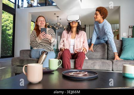 Various female friends using smartphones and VR headsets are sitting on a corner sofa in the living room. Modern, collaboration, Stock Photo