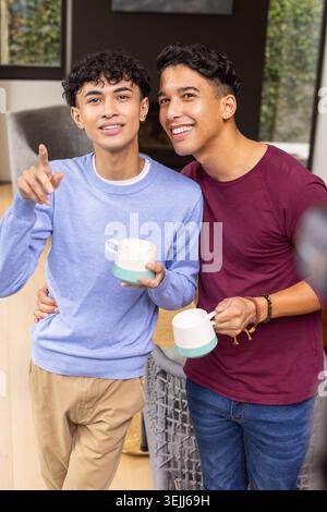 Hispanic male friends standing in living room holding mugs and pointing through window. Candid, cozy, contemporary, friendship, relaxation, domestic, Stock Photo