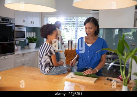 Diverse family slicing cucumber on wooden cutting board and washing produce at sink in home kitchen. Family, culinary, contemporary, lifestyle, domest Stock Photo