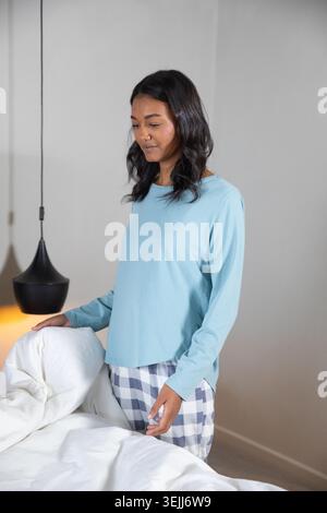 African American woman in pyjamas smoothing bedspread next to bed with lamp in bedroom, open space. Cosiness, relaxation, sereni Stock Photo