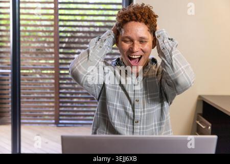 Man sitting at a bright desk in an office under sliding doors leading to the terrace, celebrating his success on his laptop. Suc Stock Photo