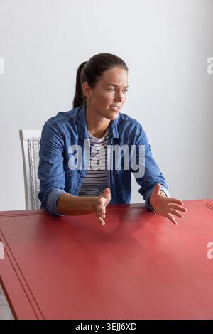 Woman gesturing with both hands and pointing to a red table in the room on a chair with a woven backrest. Professional, communic Stock Photo