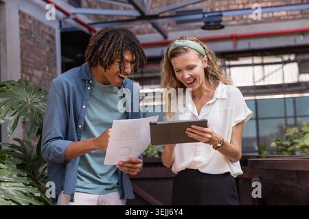 Diverse employees in business attire review data on tablets and paper maps in a loft office. Collaboration, teamwork, analytics, Stock Photo