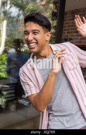 Adult man poses in front of a glass door and gestures towards the reflection of brick walls, wooden floors and foliage in his ho Stock Photo