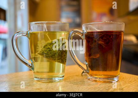 Two transparent glass mugs with green and black tea from pyramids tea bags standing on a wooden table in a cafe. Stock Photo