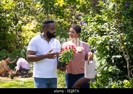 Diverse friends chatting while tending potted red plant and using white watering can in backyard. Gardening, outdoor, lush, leisure, vibrant, communit Stock Photo