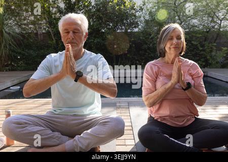 Senior couple practicing meditation sitting on deck beside pool with yoga mats and water bottle. Wellness, relaxation, tranquility, mindfulness, outdo Stock Photo