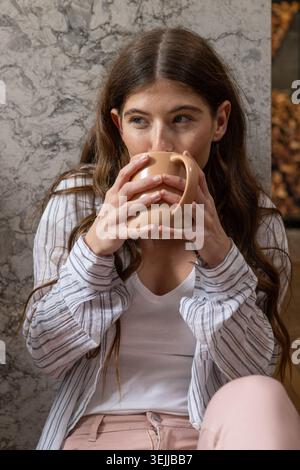 Woman in her twenties sitting against marble wall holding beige ceramic mug, gazing left. Cozy, relaxation, home interior, minim Stock Photo