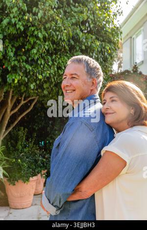 Senior couple standing on backyard patio hugging near terracotta pots, potted shrubs, window vines. Outdoor, relaxation, rustic, harmony, lifestyle, n Stock Photo