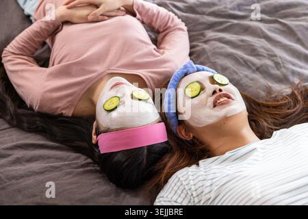 Asian mother and daughter resting on bed wearing facial masks, cucumber slices and headbands. Skincare, relaxation, selfcare, home spa, beauty ritual, Stock Photo