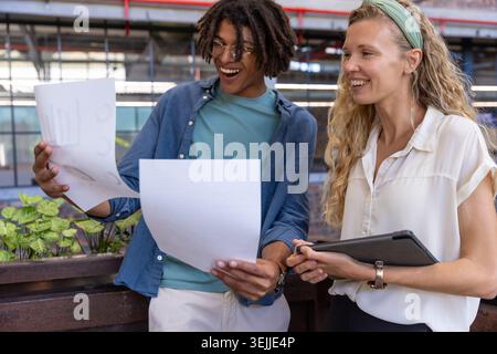 Diverse coworkers analyzing chart sheets and tablet in office atrium with planter box. Collaboration, innovation, modernity, pro Stock Photo