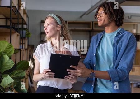 Diverse coworkers pointing at clipboard while discussing plan in shared office near potted plant. Collaboration, teamwork, moder Stock Photo