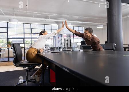 Diverse coworkers in business attire high fiving over desk at modern office with monitor and laptop. Collaboration, teamwork, productivity, corporate, Stock Photo