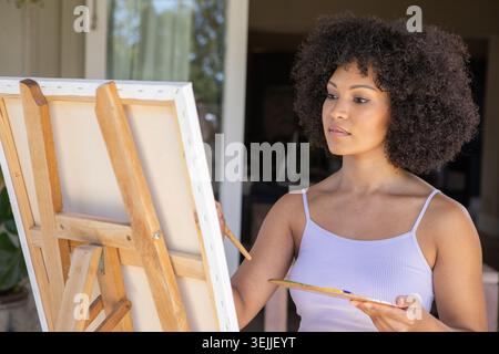 African American woman painting on canvas at covered patio using wooden easel, holding paintbrush. Art, creativity, outdoors, serene, artisan, modern, Stock Photo