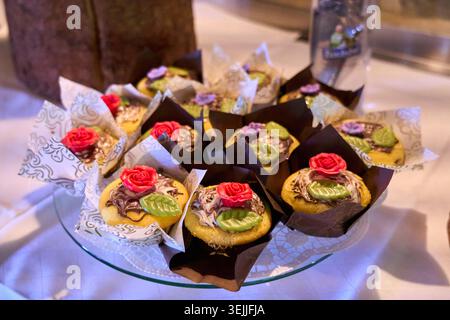 Assortment of decorated cupcakes with buttercream roses and green leaves on a glass platter, served at a dessert buffet. Stock Photo