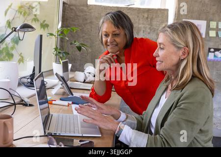 Diverse senior female colleagues collaborating over laptop and VR headset at modern office desk. Collaboration, teamwork, modernity, innovation, works Stock Photo