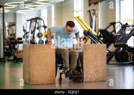 Confident man athlete in wheelchair doing push-ups strengthening exercise on wooden cubes in gym. Stock Photo