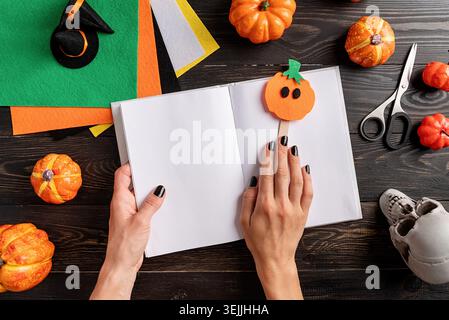 Happy Halloween concept. Making Halloween pumpkin bookmark craft. Woman hands with black nails holding pupmkin bookmark in a blank opened book. Stock Photo