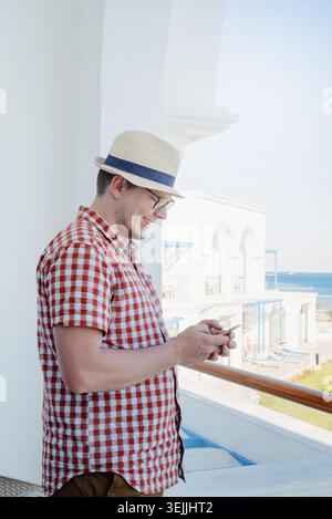 Handsome Caucasian young man standing on balcony of hotel, using mobile phone Stock Photo