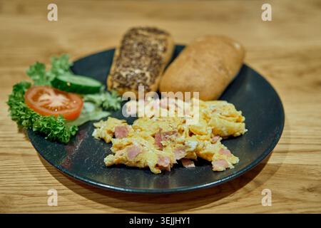 Delicious breakfast of scrambled eggs with ham, fresh buns, and sliced vegetables served on a black plate. Stock Photo