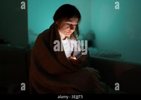 Sleepy woman sitting on couch using smartphone late at night, can not sleep. Insomnia, addiction. Stock Photo