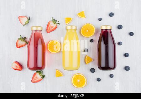 Organic yellow orange, pink strawberry and violet blueberry juices in glass bottles with fruit ingredients on white wood board, Stock Photo