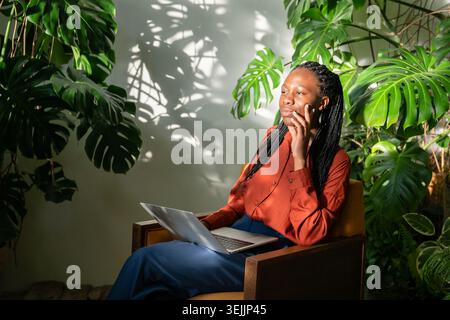 Thoughtful calm African American woman relaxing in chair with laptop surrounded by tropical plants. Stock Photo
