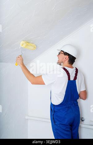 Young man in blue overalls and white hard hat painting wall into white color with a roller Stock Photo