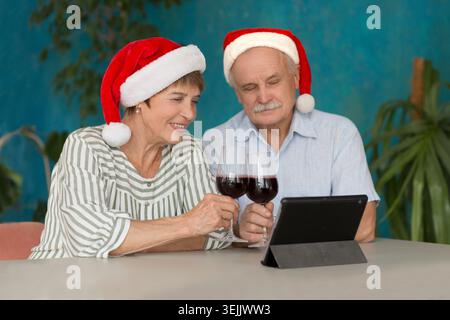 Elderly couple in a Santas hat   drinking wine and using laptop and having video call with friends or relatives at home. Christm Stock Photo