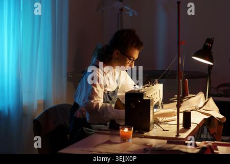 Busy designer woman working late at night in atelier with sewing machine, fabric and paper patterns Stock Photo