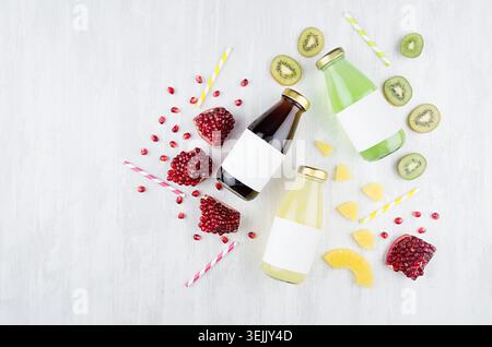 Fresh yellow pineapple, green kiwi, pomegranate juices in glass bottles with blank label, ingredients on white wood board, mock Stock Photo