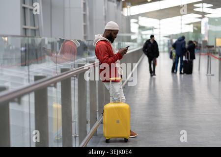 Thoughtful African American traveler man using smartphone at airport terminal, travelling with luggage bag. Passenger guy waiting her flight, checks t Stock Photo